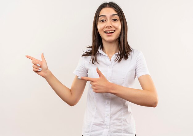 Giovane ragazza in camicia bianca che punta con il dito indice al lato sorridente in piedi sopra il muro bianco