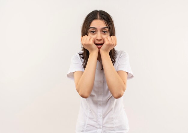 Giovane ragazza in camicia bianca che sembra stressata e nervosa in piedi sopra il muro bianco