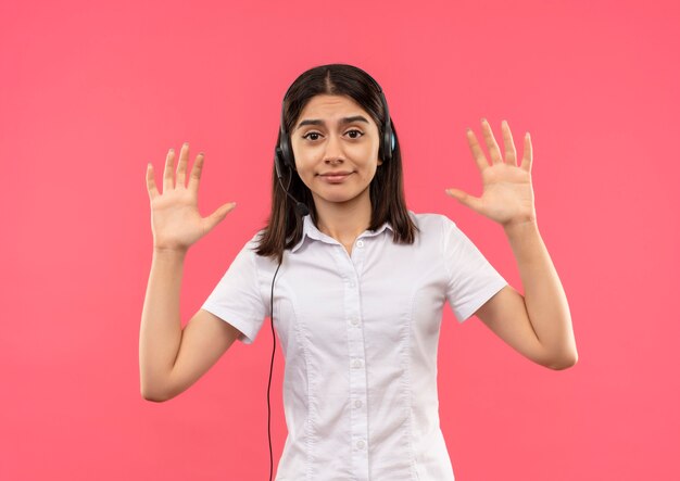 Giovane ragazza in camicia bianca e cuffie, alzando le mani in segno di resa cercando confuso in piedi sopra il muro rosa