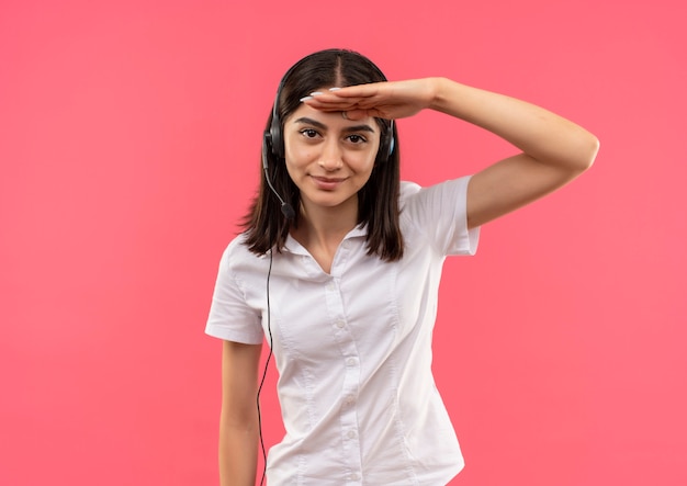 Giovane ragazza in camicia bianca e cuffie, guardando lontano con la mano sulla testa in piedi sul muro rosa