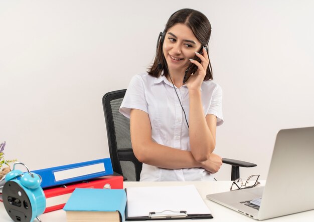 Giovane ragazza in camicia bianca e cuffie, parlando al telefono cellulare sorridente seduto al tavolo con cartelle e laptop sul muro bianco