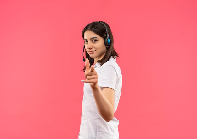 Giovane ragazza in camicia bianca e cuffie, puntando il dito verso la parte anteriore sorridente fiducioso in piedi oltre il muro rosa