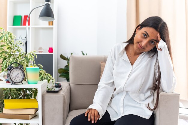 Giovane ragazza in camicia bianca e pantaloni neri che guarda l'obbiettivo sorridente felice e positivo seduto sulla sedia in soggiorno luminoso