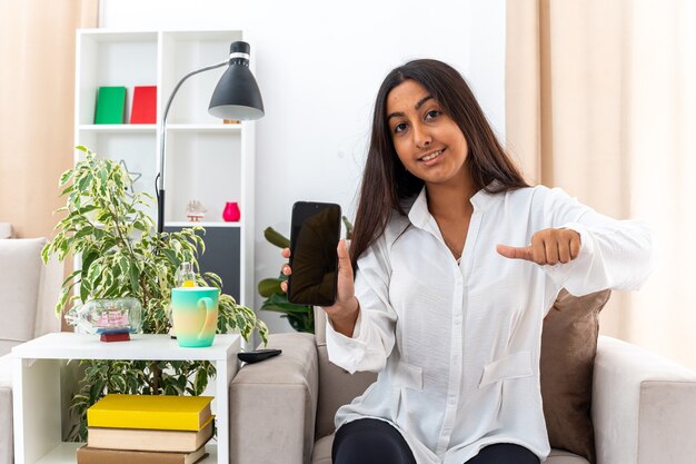 Giovane ragazza in camicia bianca e pantaloni neri che tiene in mano uno smartphone che mostra i pollici in su felice e allegra seduta sulla sedia in un soggiorno luminoso