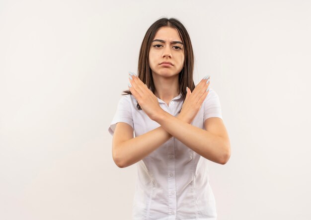 Giovane ragazza in camicia bianca guardando in avanti con la faccia seria che fa smettere di cantare attraversando le mani in piedi sul muro bianco