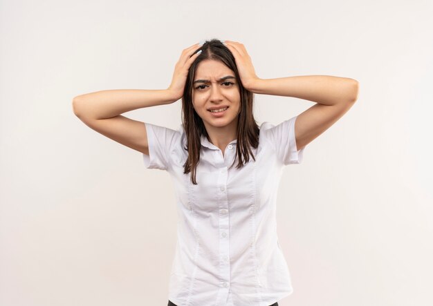 Giovane ragazza in camicia bianca guardando in avanti confuso e deluso tenendo la sua testa con le mani in piedi sul muro bianco