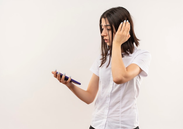 Giovane ragazza in camicia bianca guardando lo schermo del suo telefono cellulare confuso in piedi sopra il muro bianco
