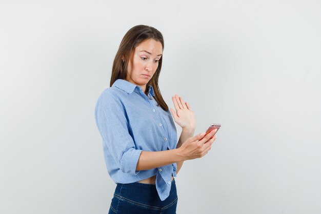 Giovane ragazza in camicia blu, pantaloni agitando la mano, tenendo lo smartphone e guardando confuso
