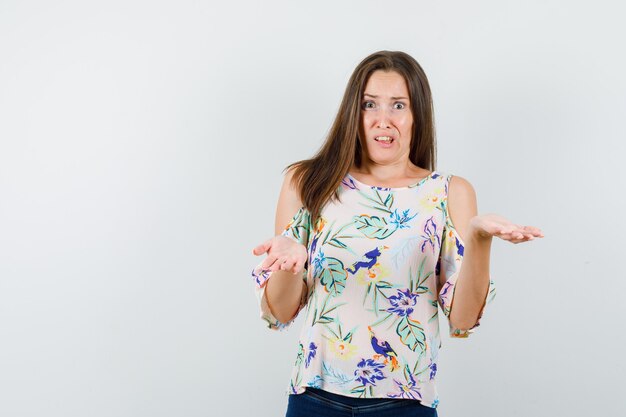 Giovane ragazza in camicia, jeans alzando le palme nel gesto interrogativo e guardando infastidito, vista frontale.