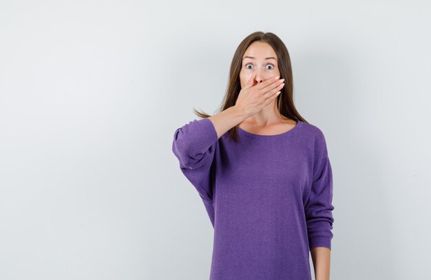 Giovane ragazza in camicia viola che copre la bocca con la mano e guardando spaventato, vista frontale.