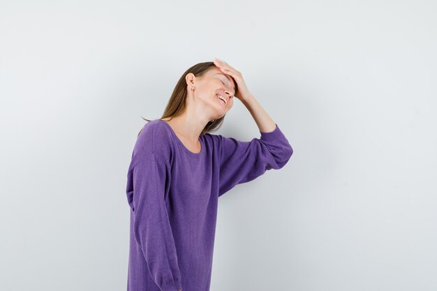 Giovane ragazza in camicia viola tenendo la mano sulla fronte e guardando felice, vista frontale.