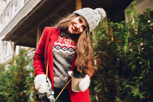 Giovane ragazza in cappotto rosso che cammina sulla strada con il caffè per andare. Indossa guanti bianchi, sorridendo.