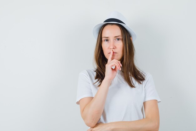Giovane ragazza in maglietta bianca, cappello che mostra gesto di silenzio e guardando paziente, vista frontale.