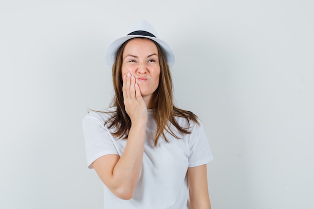 Giovane ragazza in maglietta bianca, cappello che soffre di mal di denti e che sembra a disagio, vista frontale.