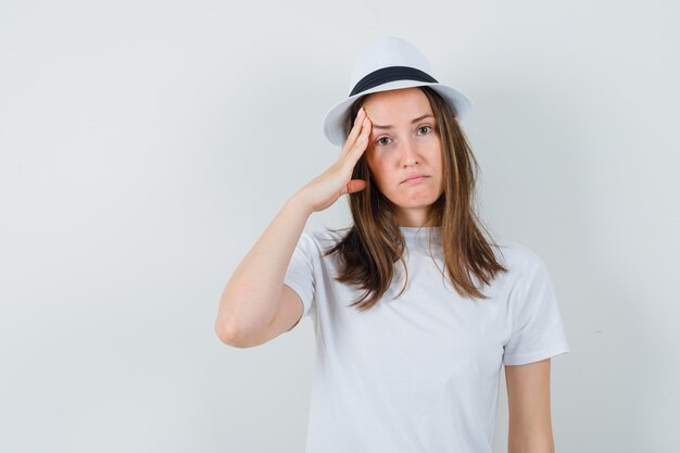 Giovane ragazza in maglietta bianca, cappello che tira la pelle sulle tempie e sembra triste, vista frontale.