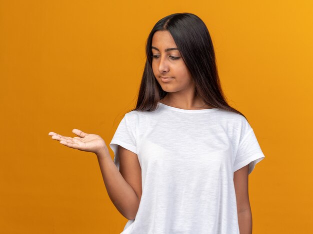 Giovane ragazza in maglietta bianca che guarda da parte con espressione sicura che presenta lo spazio della copia con il braccio della mano in piedi su sfondo arancione