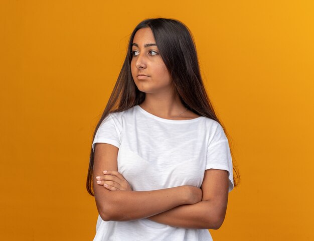 Giovane ragazza in maglietta bianca che guarda da parte con una faccia seria con le braccia incrociate in piedi sopra l'arancia