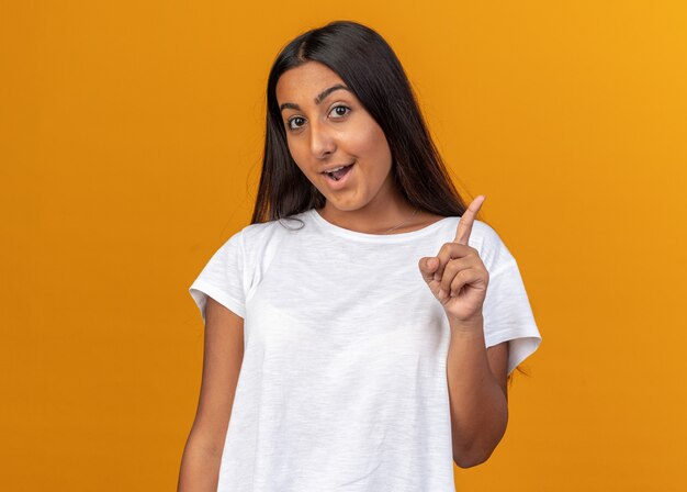 Giovane ragazza in maglietta bianca che guarda la telecamera con un sorriso sul viso intelligente che mostra il dito indice che ha una grande idea in piedi sopra l'arancia