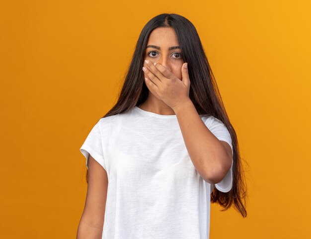 Giovane ragazza in maglietta bianca che guarda la telecamera scioccata che copre la bocca con la mano in piedi su uno sfondo arancione