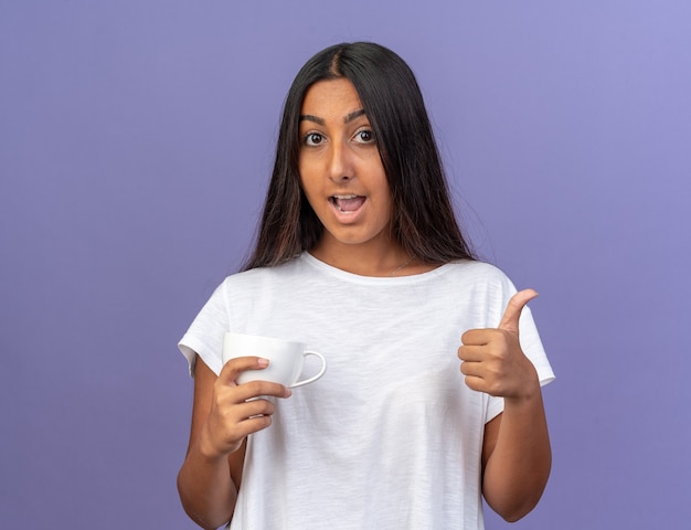 Giovane ragazza in maglietta bianca che guarda la telecamera sorridendo allegramente mostrando i pollici in piedi sopra il blu