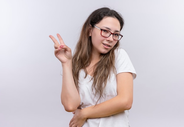 Giovane ragazza in maglietta bianca che sorride allegramente mostrando il segno di vittoria