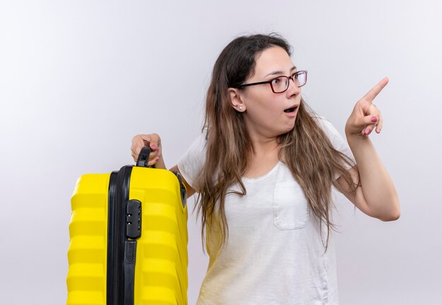 Giovane ragazza in maglietta bianca che tiene la valigia di viaggio che osserva da parte sorpreso e stupito che indica con il dito indice a qualcosa