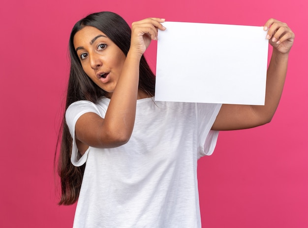 Giovane ragazza in maglietta bianca che tiene un foglio di carta bianco bianco che guarda la telecamera sorpresa e stupita in piedi su sfondo rosa