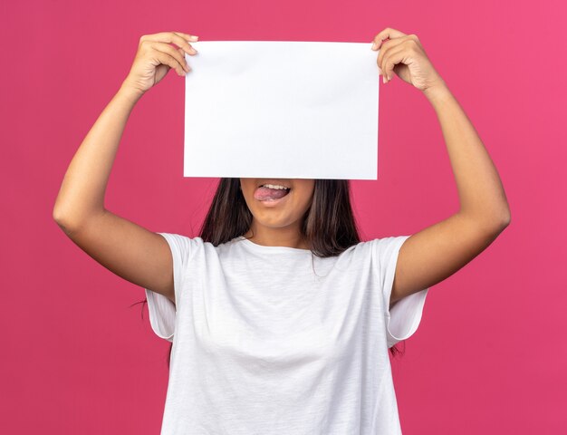 Giovane ragazza in maglietta bianca che tiene un foglio di carta bianco bianco davanti al viso sorridente in piedi sopra il rosa