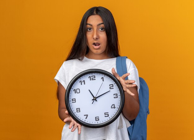 Giovane ragazza in maglietta bianca con orologio da parete che guarda la telecamera stupita e sorpresa in piedi su sfondo arancione