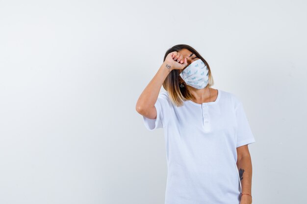 Giovane ragazza in maglietta bianca e maschera strofinando gli occhi con il pugno e guardando stanco, vista frontale.