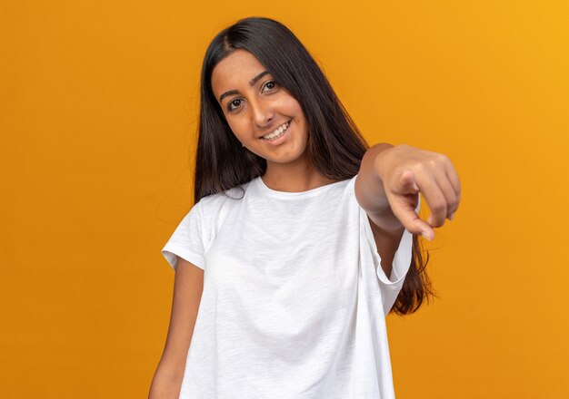 Giovane ragazza in maglietta bianca felice e positiva che punta con il dito indice alla telecamera sorridendo allegramente in piedi su sfondo arancione