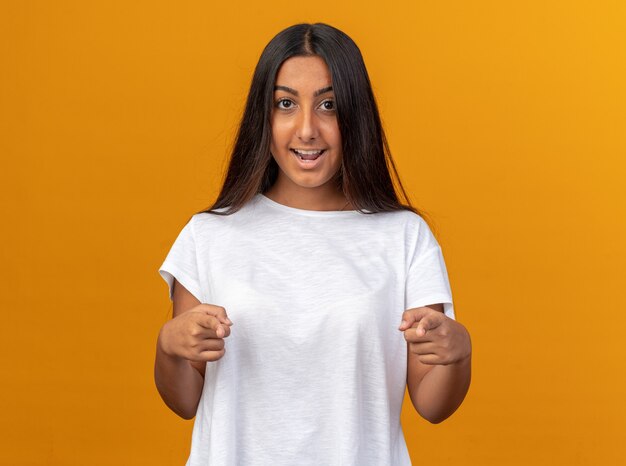 Giovane ragazza in maglietta bianca felice e positiva che punta con il dito indice alla telecamera sorridendo allegramente in piedi su sfondo arancione