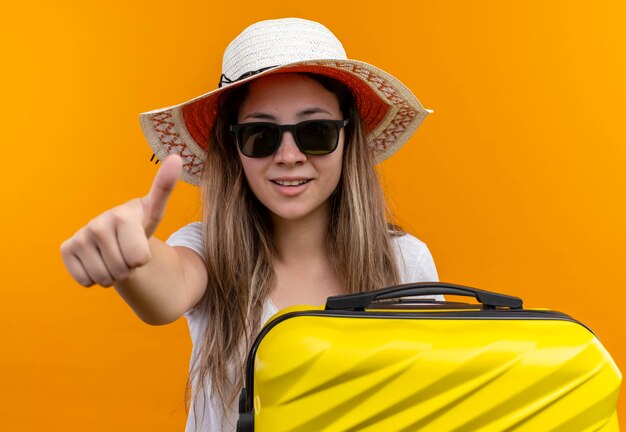 Giovane ragazza in maglietta bianca in cappello estivo che tiene la valigia di viaggio sorridente che mostra i pollici in su