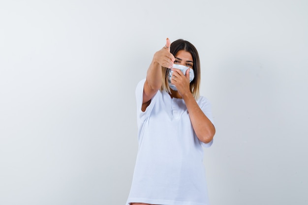 Giovane ragazza in maglietta bianca, maschera che mostra il gesto della pistola, che copre la bocca con la mano e sembra sicura, vista frontale.