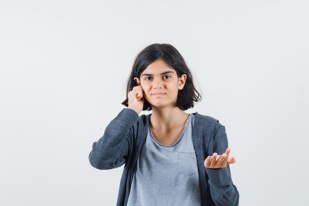 Giovane ragazza in maglietta grigio chiaro e felpa con cappuccio con zip grigio scuro che allunga la mano verso la telecamera mentre riceve qualcosa e mette la mano sull'orecchio e sembra carina,