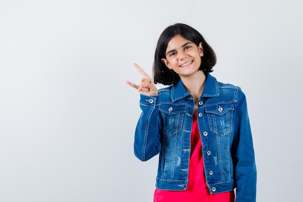 Giovane ragazza in maglietta rossa e giacca di jeans che mostra il gesto del rock n roll e sembra carina, vista frontale.