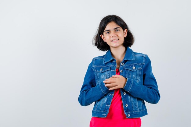 Giovane ragazza in maglietta rossa e giacca di jeans che si sfrega le mani e sembra allegra, vista frontale.