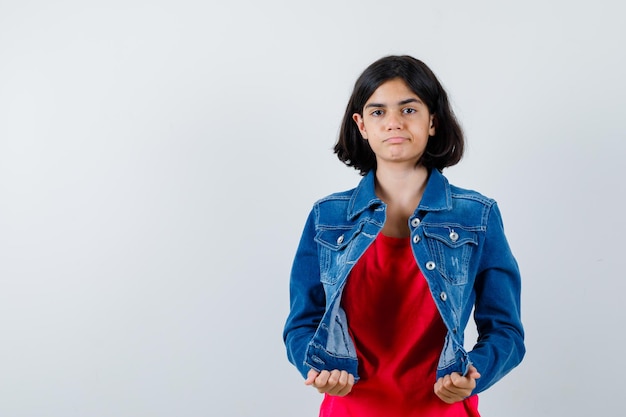 Giovane ragazza in maglietta rossa e giacca di jeans che tiene la giacca con le mani e sembra seria, vista frontale.