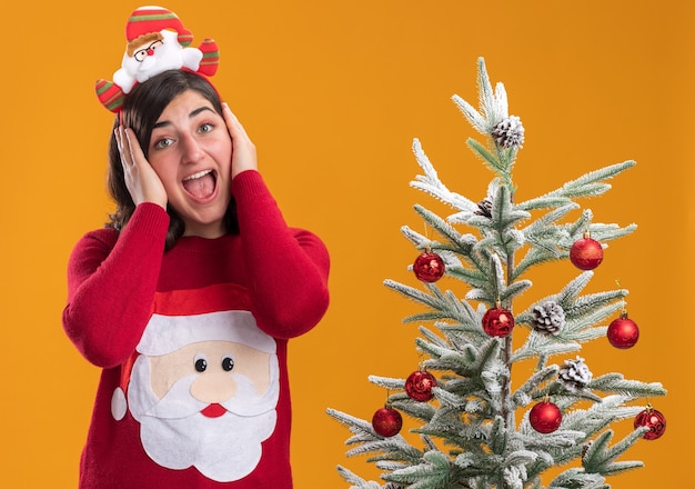 Giovane ragazza in maglione di Natale che indossa la fascia divertente pazzo felice che copre le orecchie che grida in piedi accanto a un albero di Natale oltre la parete arancione