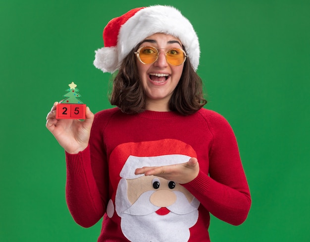 Giovane ragazza in maglione di Natale che indossa un cappello da Babbo Natale e occhiali tenendo i cubi giocattolo con il numero venticinque sorridendo allegramente presentando con il braccio in piedi sopra la parete verde