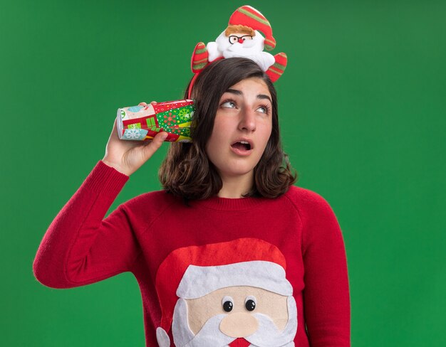 Giovane ragazza in maglione di Natale che porta il cappello della Santa che tiene il bicchiere di carta colorato sopra l'orecchio cercando di ascoltare qualcosa in piedi sopra la parete verde