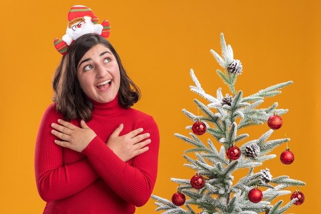 Giovane ragazza in maglione di Natale indossando la fascia divertente accanto a un albero di Natale su sfondo arancione