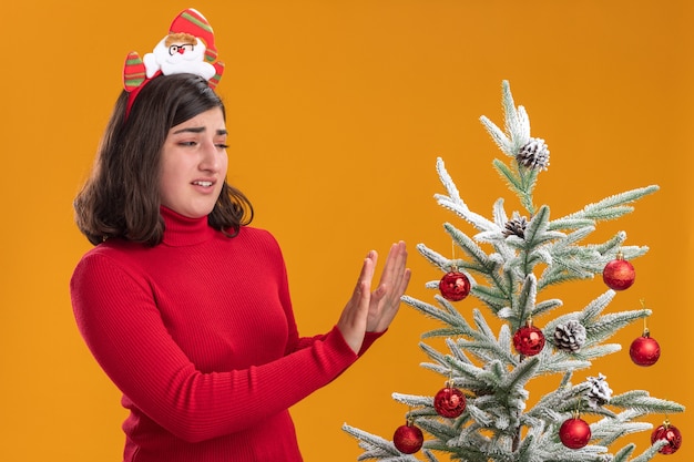 Giovane ragazza in maglione di Natale indossando la fascia divertente accanto a un albero di Natale su sfondo arancione