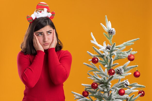 Giovane ragazza in maglione di Natale indossando la fascia divertente che osserva da parte confusa e molto ansiosa accanto a un albero di Natale su sfondo arancione