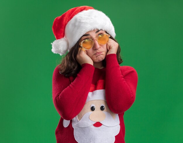 Giovane ragazza in maglione di Natale santa cappello e occhiali guardando da parte con espressione triste in piedi sopra la parete verde