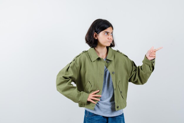 Giovane ragazza in maglione grigio, giacca color kaki, pantaloni di jeans che puntano a destra mentre si tiene la mano sulla vita e sembra carina, vista frontale.