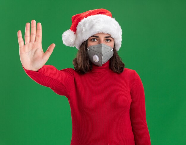 Giovane ragazza in maglione rosso e cappello da Babbo Natale che indossa la maschera protettiva per il viso con la faccia seria che fa il gesto di arresto con la mano che sta sopra la parete verde