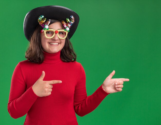 Giovane ragazza in maglione rosso indossando occhiali divertenti e cappello nero guardando sorridente della fotocamera