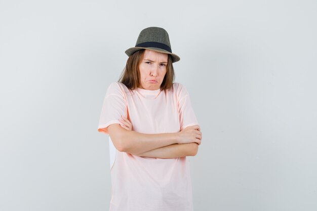 Giovane ragazza in piedi con le braccia incrociate in cappello t-shirt rosa e guardando offeso