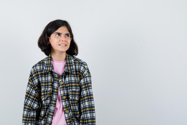 Giovane ragazza in piedi dritta, guardando lontano e posa alla macchina fotografica in camicia a quadri e t-shirt rosa e sembra carina. vista frontale.